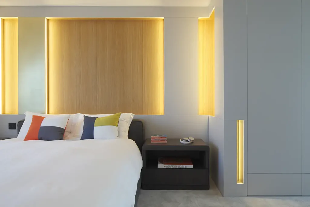 Modern bedroom with a bed featuring multicolored pillows, a wooden accent wall with ambient lighting, and a dark bedside table holding books. The design seamlessly merges comfort and style, showcasing the best in contemporary architecture. Map Projects