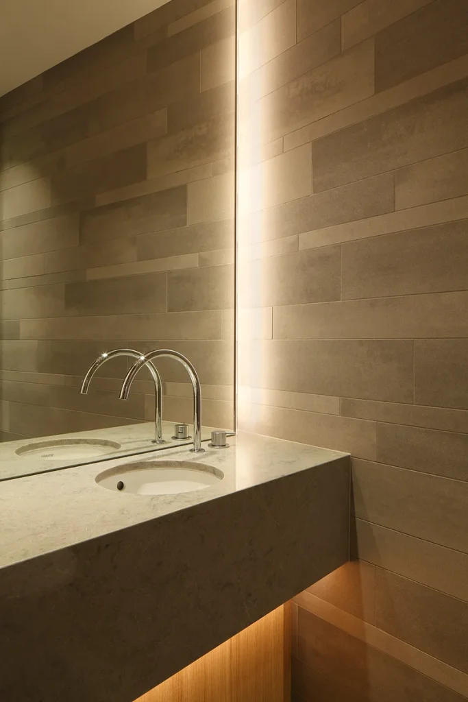 Modern bathroom with a minimalist countertop and dual faucets, showcasing exquisite design. Set against a textured beige tile wall with ambient lighting, it reflects sleek architecture. Map Projects