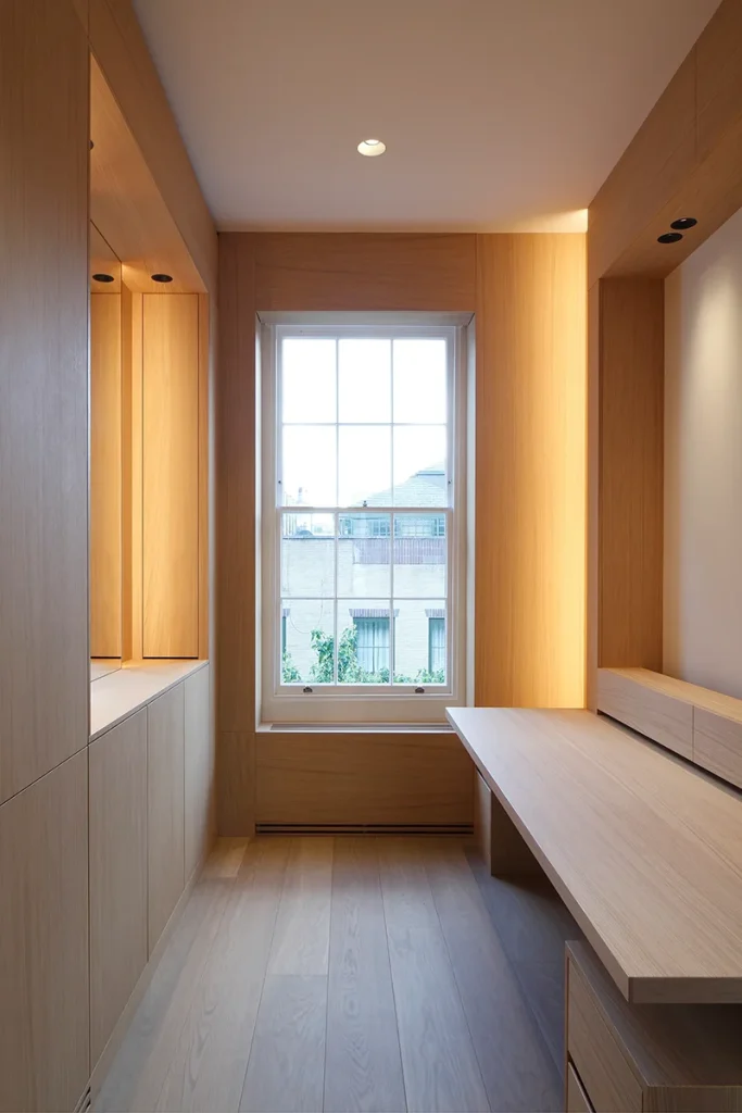 Minimalist room with light wood furniture, including shelves and a desk. The architectural design features a large window with vertical bars and a view of neighboring buildings. A ceiling light elegantly illuminates the space. Map Projects