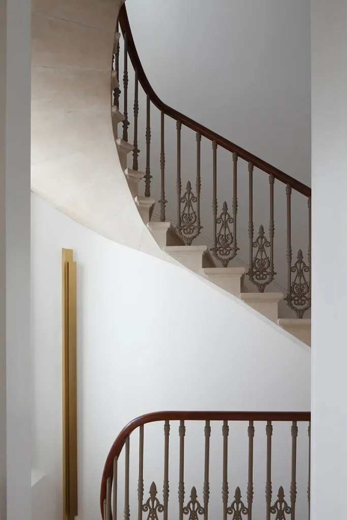 The design of the spiraling staircase, with its ornate metal railings and polished wooden handrails, adds a touch of elegance to the white-walled architectural interior. Map Projects