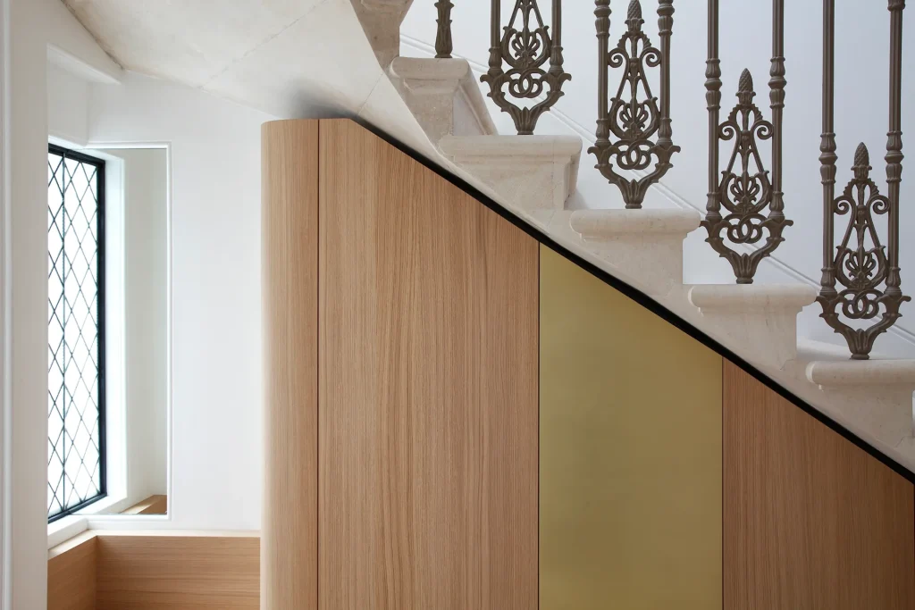 A modern staircase with architectural elegance features a decorative metal railing and wooden paneling underneath. On the left, a window with diamond-patterned glass enhances the design. Map Projects