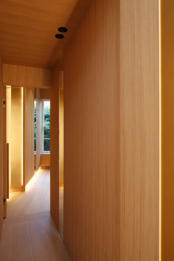 A modern hallway showcases exquisite architecture with wooden walls and floors, enhanced by sleek recessed lighting. The design is perfected by a window at the end offering a serene view of greenery. Map Projects