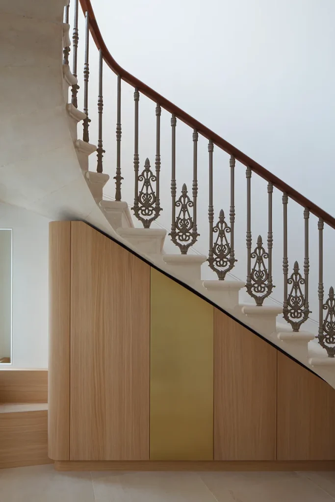 The architecture features a staircase with an ornate metal railing that gracefully curves above light wood paneling, accented with gold against a pristine white wall, showcasing exquisite design. Map Projects