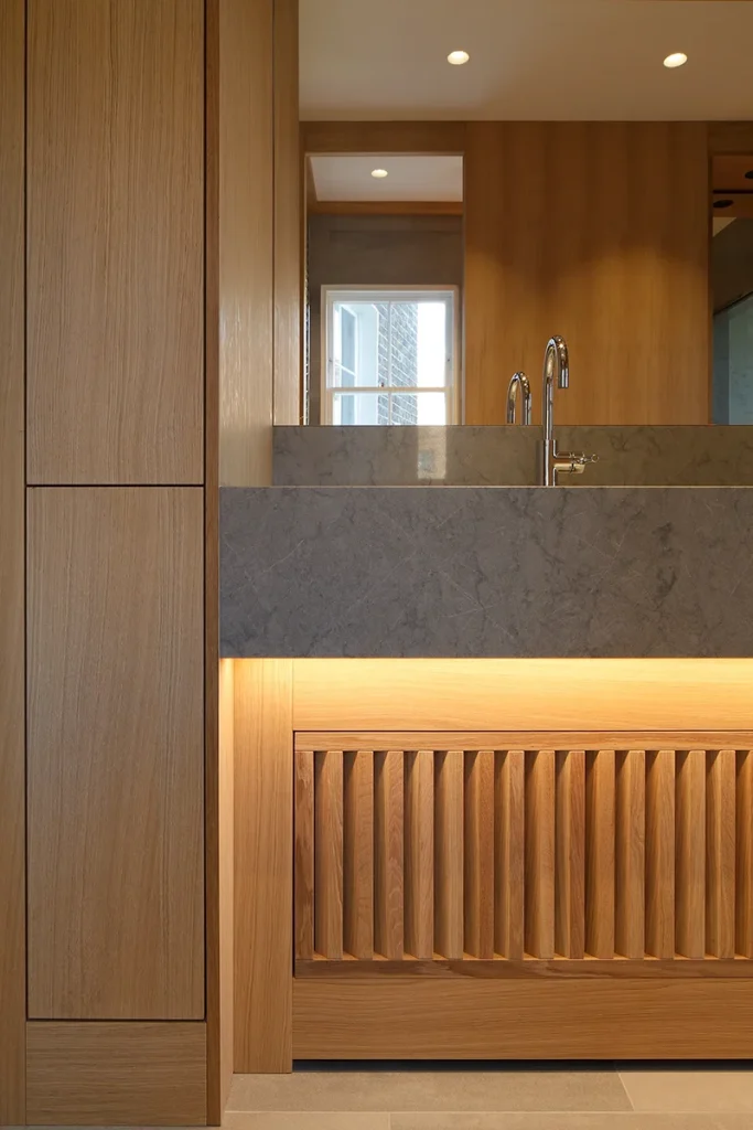 Modern bathroom vanity with a gray stone countertop, chrome faucet, and vertical wooden slat cabinet, showcasing exquisite design. Warm accent lighting beneath enhances the sophisticated architecture of this stylish space. Map Projects