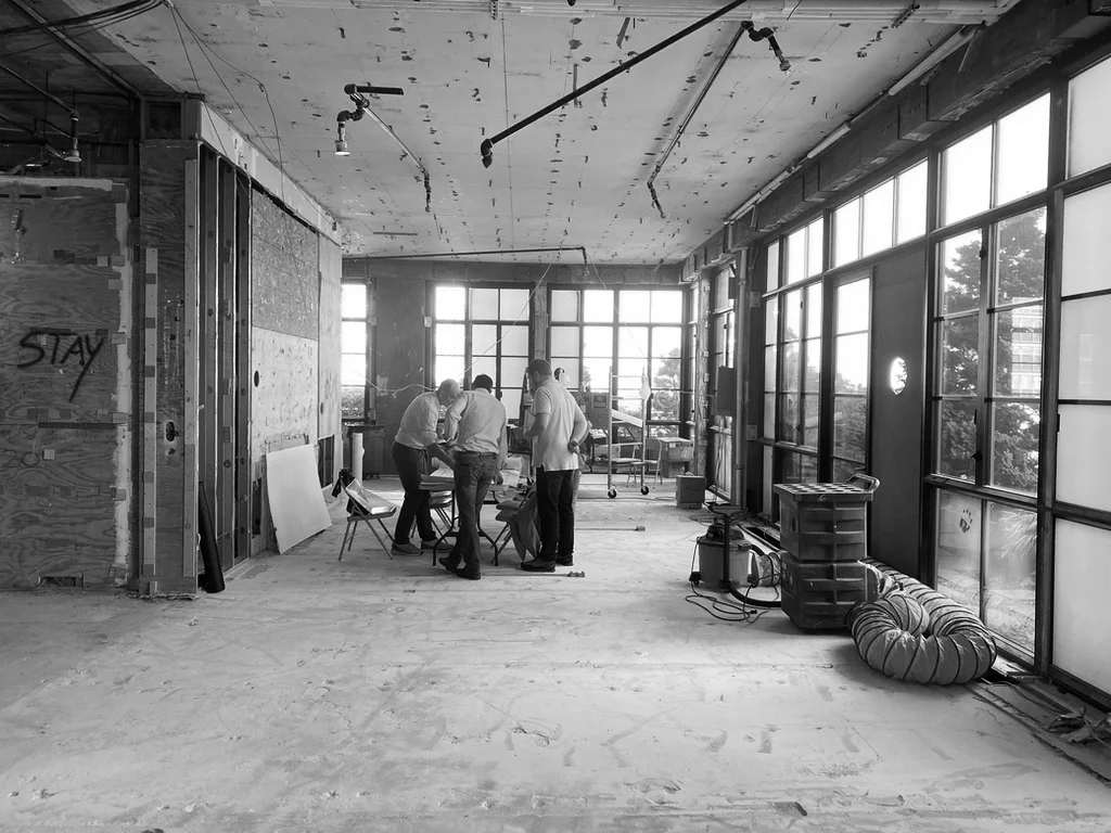 Three people stand around a table discussing design strategies in a partially renovated room filled with exposed wiring and construction materials, highlighting the essence of architecture in progress. Map Projects