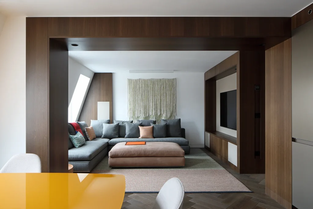Modern living room with a design-focused gray sectional sofa, a large ottoman, and a wall-mounted TV. A yellow-tinted glass table sits in the foreground. Dark wooden paneling frames the space, adding architectural elegance. Map Projects