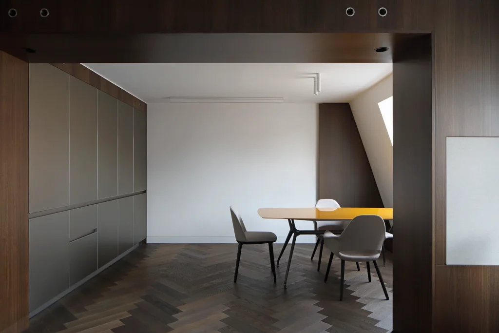 Modern dining room featuring a sleek yellow table, four chairs, and gray cabinetry, all showcasing impeccable design. Herringbone wood flooring complements the neutral walls, while natural light enhances the minimalist architecture. Map Projects