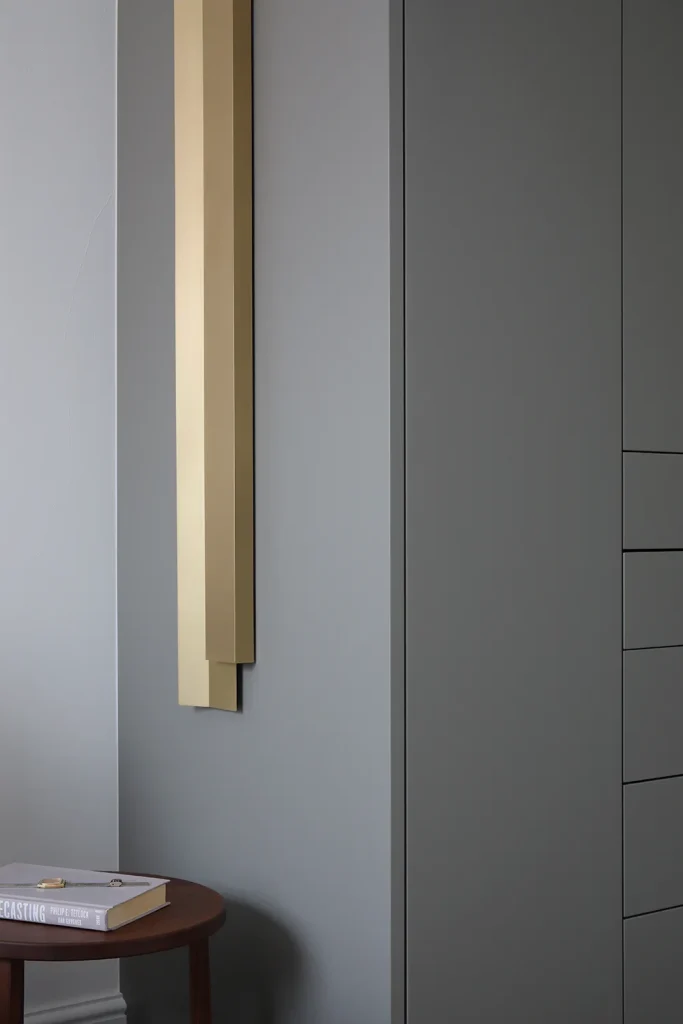 Minimalist interior design with a vertical gold wall art piece adorning a gray wall, beside a wooden stool with a book. Map Projects