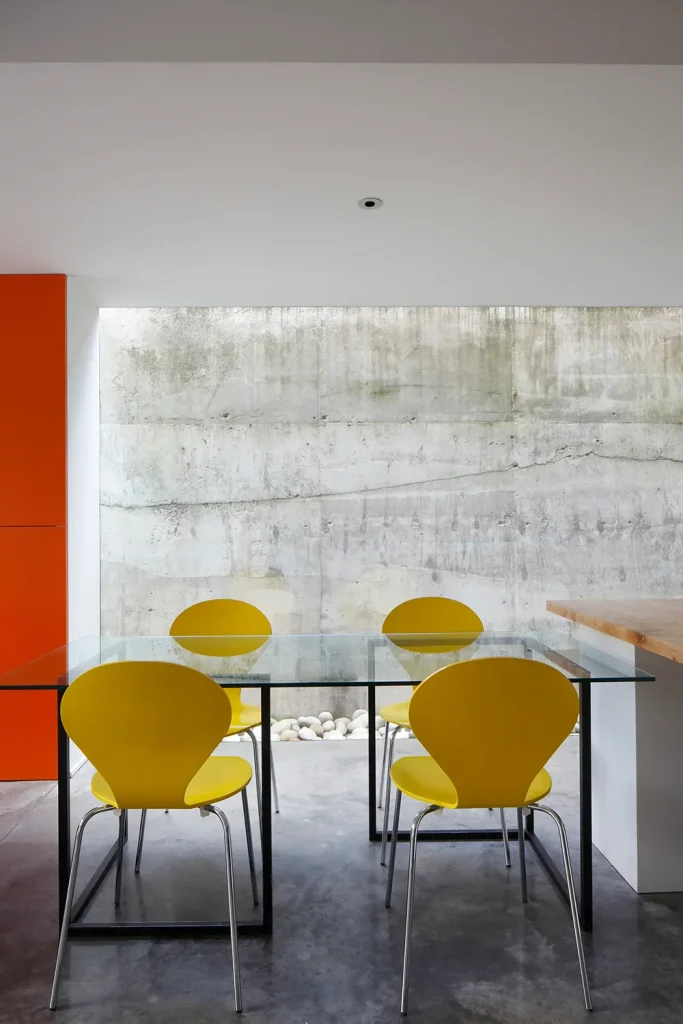 A modern dining area showcases sleek architecture with a glass table and four yellow chairs on a gray concrete floor, seamlessly blending into the stylish design of the large concrete wall in the background. Map Projects