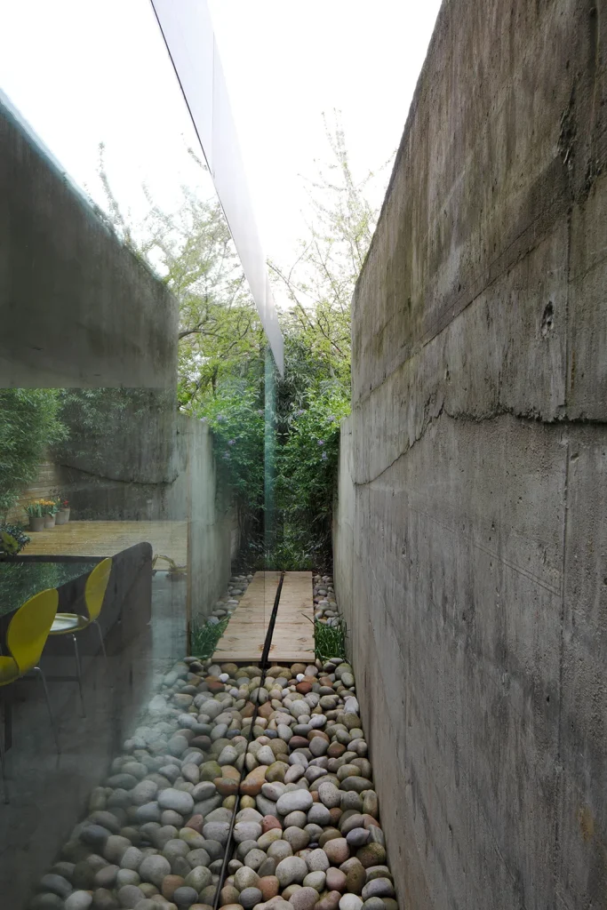 A narrow outdoor path with a wooden walkway, showcasing exquisite design, runs between a concrete wall and a glass wall, reflecting chairs and foliage in an architecture marvel flanked by pebbles. Map Projects