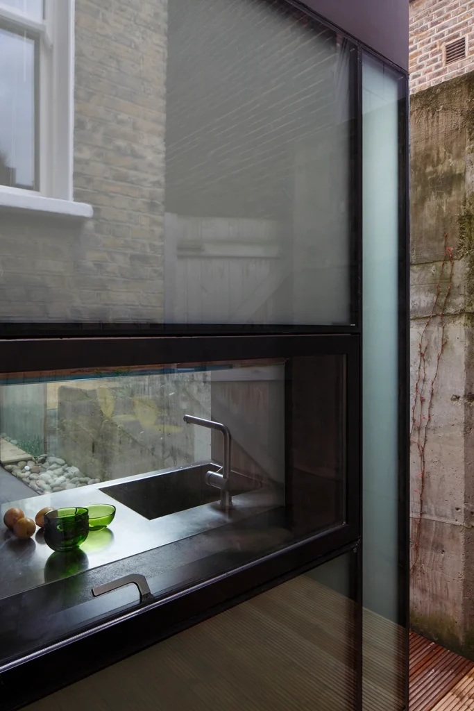 A modern kitchen sink area showcases exquisite design, featuring a double window view and a green glass bottle on the counter, complemented by the striking architecture of a brick wall outside. Map Projects