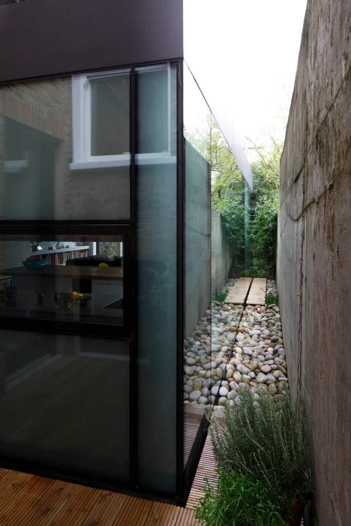 Narrow outdoor pathway with smooth stones and lush greenery beside a modern glass-walled building, showcasing exquisite architecture. Sleek concrete wall on the right adds to the contemporary design. Map Projects