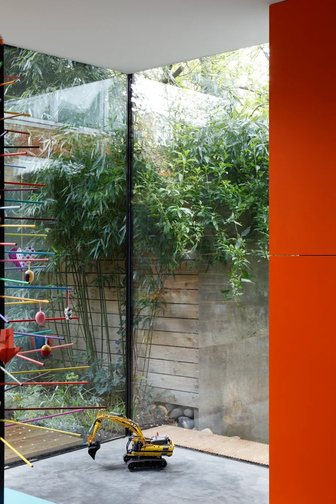 A model excavator sits on a concrete floor in a modern room with large glass windows, seamlessly blending design and architecture. Outside, a bamboo garden and wooden fence provide contrast, while vertical orange and colorful bars decorate the interior. Map Projects