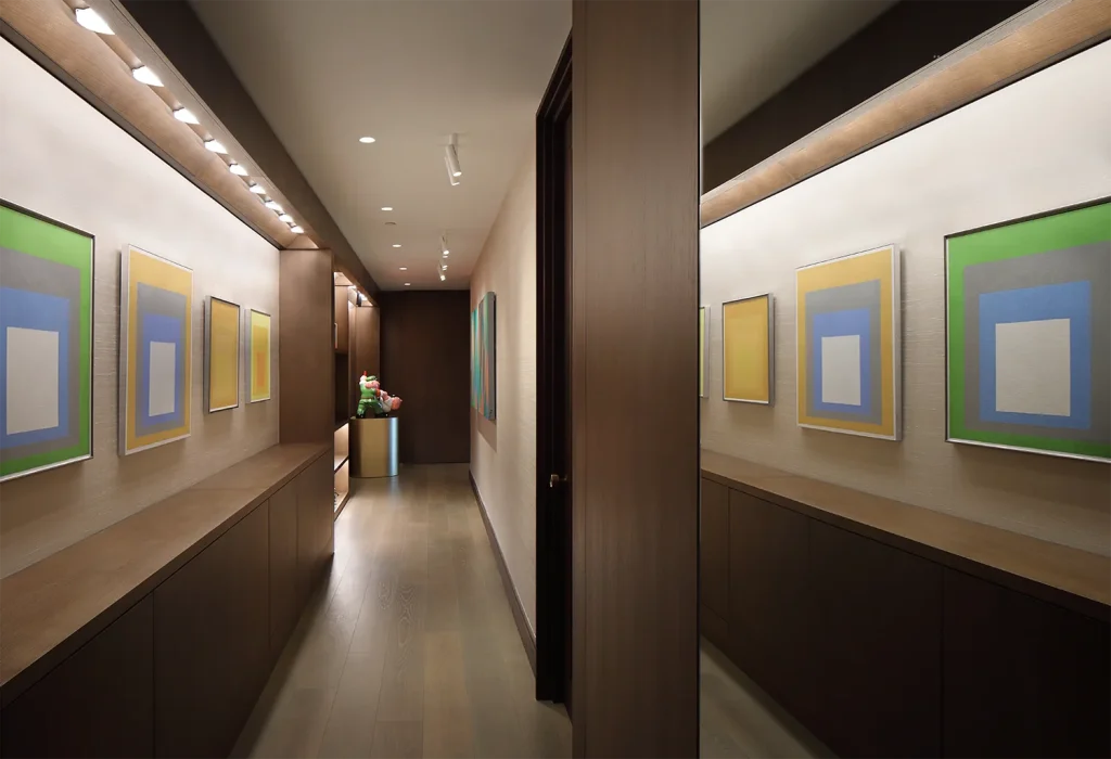 A hallway with modern artwork on both sides, wooden flooring, and warm lighting showcases exquisite design. A doorway reveals a glimpse of a room adorned with flowers at the end, completing this architectural masterpiece. Map Projects