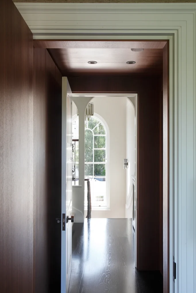 View through a wooden doorway into a bright room that showcases stunning architecture, featuring a large arched window framing the greenery outside. The modern chandelier complements the elegant design, with dark hardwood flooring throughout. Map Projects