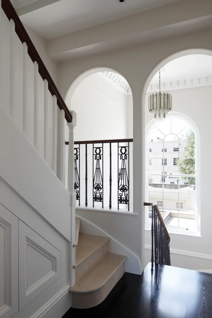 Elegant staircase with wooden steps and decorative iron railing showcases exquisite architecture, flanked by arched windows, leading to a well-lit landing with a chandelier that epitomizes refined design. Map Projects