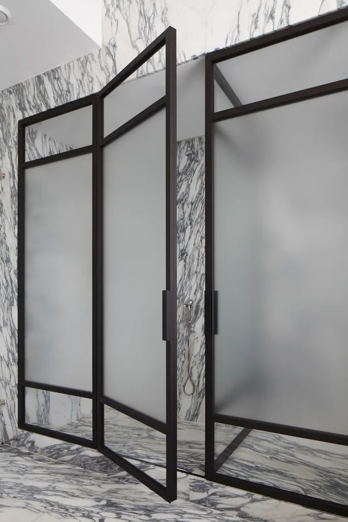 Frosted glass shower doors with black metal frames enhance the design of a marble-tiled bathroom, creating a seamless blend of contemporary style and elegant architecture. Map Projects