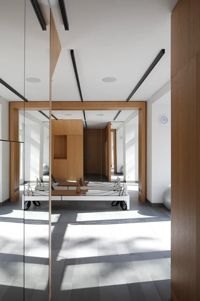 Modern fitness room with a Pilates reformer, wooden accents, and large mirrors reflecting thoughtful design. A wall clock adds functionality, while bright natural light highlights the architectural elegance of the space. Map Projects