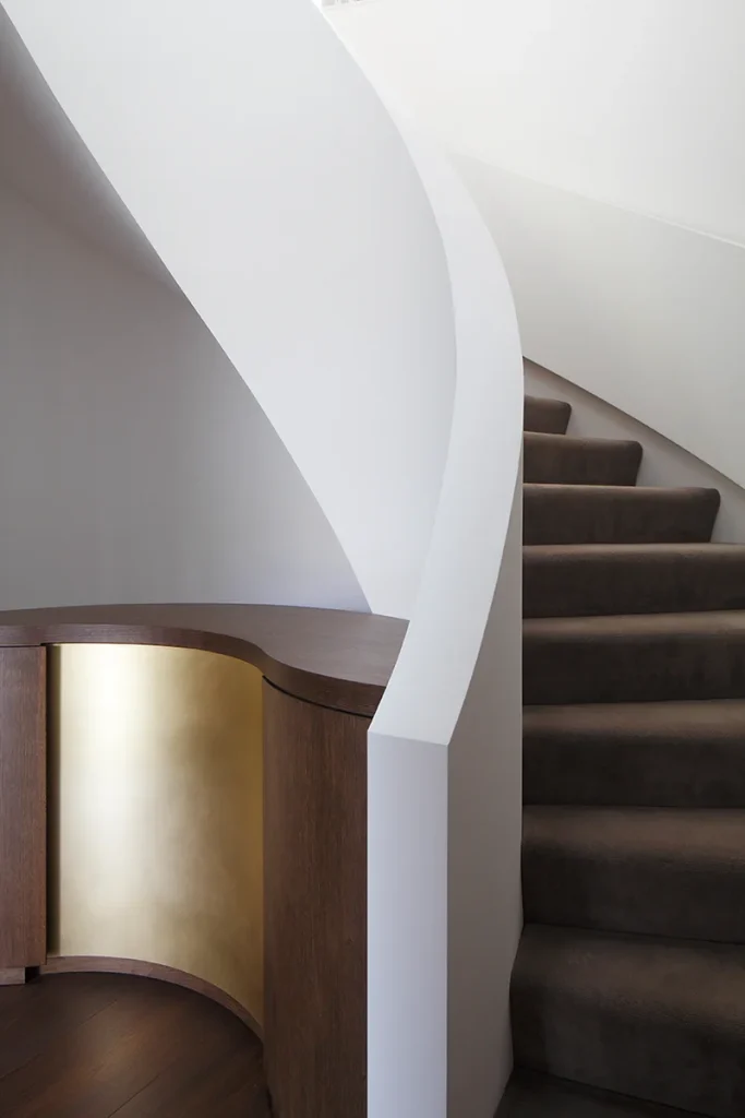 A modern interior design showcases a curved white staircase with brown carpeted steps, complemented by a gold-accented wooden cabinet at the base, highlighting exceptional architecture. Map Projects