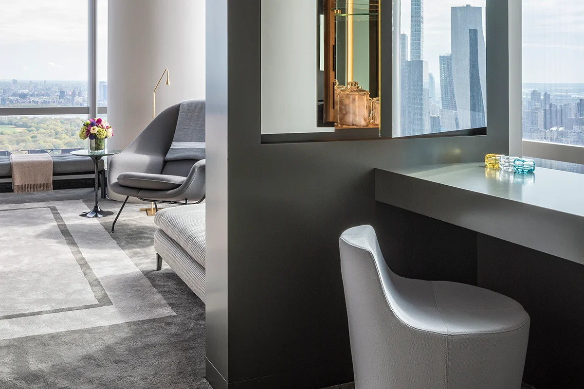 A modern hotel room featuring a sleek desk, ergonomic chair, and cozy lounge area. Large windows showcase a stunning cityscape and park view. The minimalist decor is enhanced by colorful glass items and flowers, reflecting thoughtful design elements that echo the surrounding architecture. Map Projects