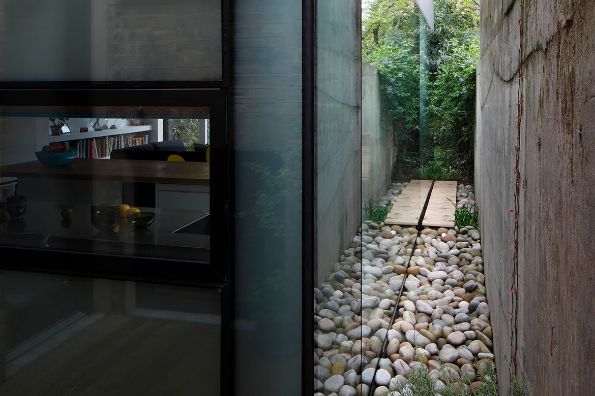 A modern interior featuring sleek design and a glass wall reveals an outdoor walkway with smooth stones and concrete on both sides, embodying contemporary architecture as it leads towards lush greenery. Map Projects