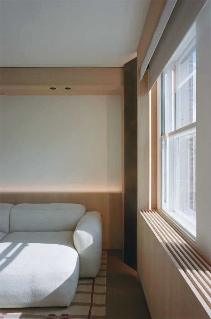 Small lounge area with custom sofa, timber shelving, and narrow window revealing textured façade